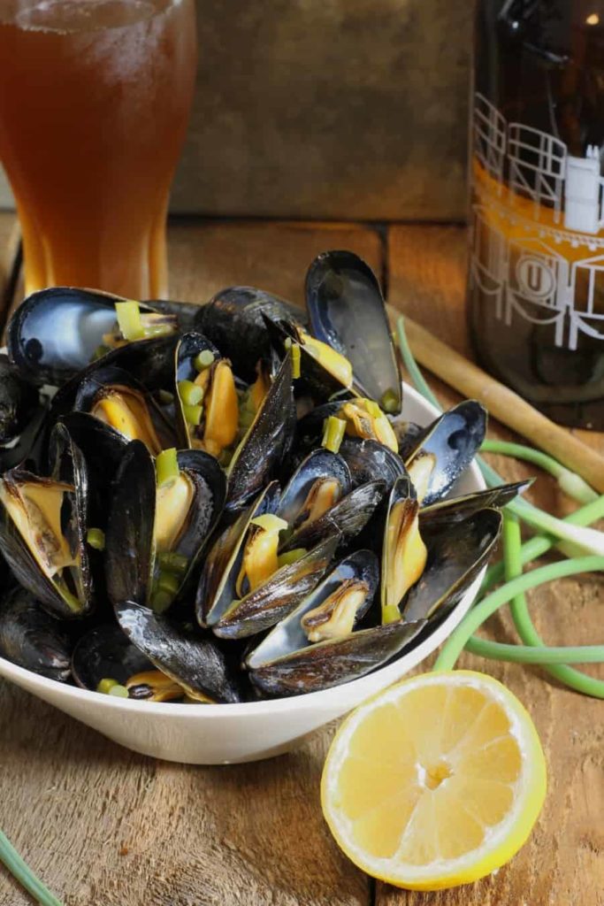 A vertical image of beer steamed mussels tossed with garlic scapes and lemon served in a white bowl