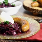 braised german red cabbage served on a fancy gold rimmed glass plate alongside sausages and mashed potatoe.