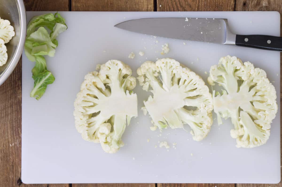 Three cauliflower steaks laying on a white cutting board  beside a chefs knife.
