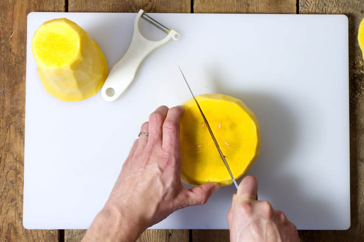 Cutting the bottom 'bulb' end of a butternut squash in half on a white cutting board.