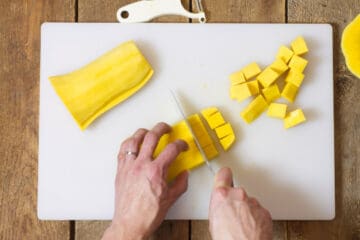 Overhead view of butternut squash being cut into chunks on a white cutting board.
