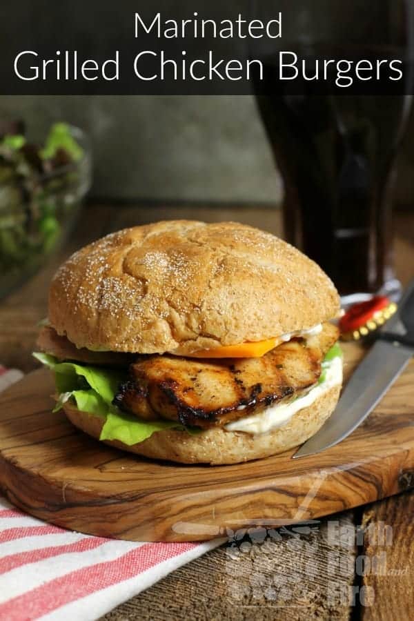a marinated and grilled chicken breast burher on a wooden serving board. a banner reading marinated grilled chicken burger is overlaid at the top of the image