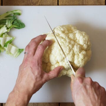 An over head shot of a head of cauliflower being cut in half on a white cutting board.