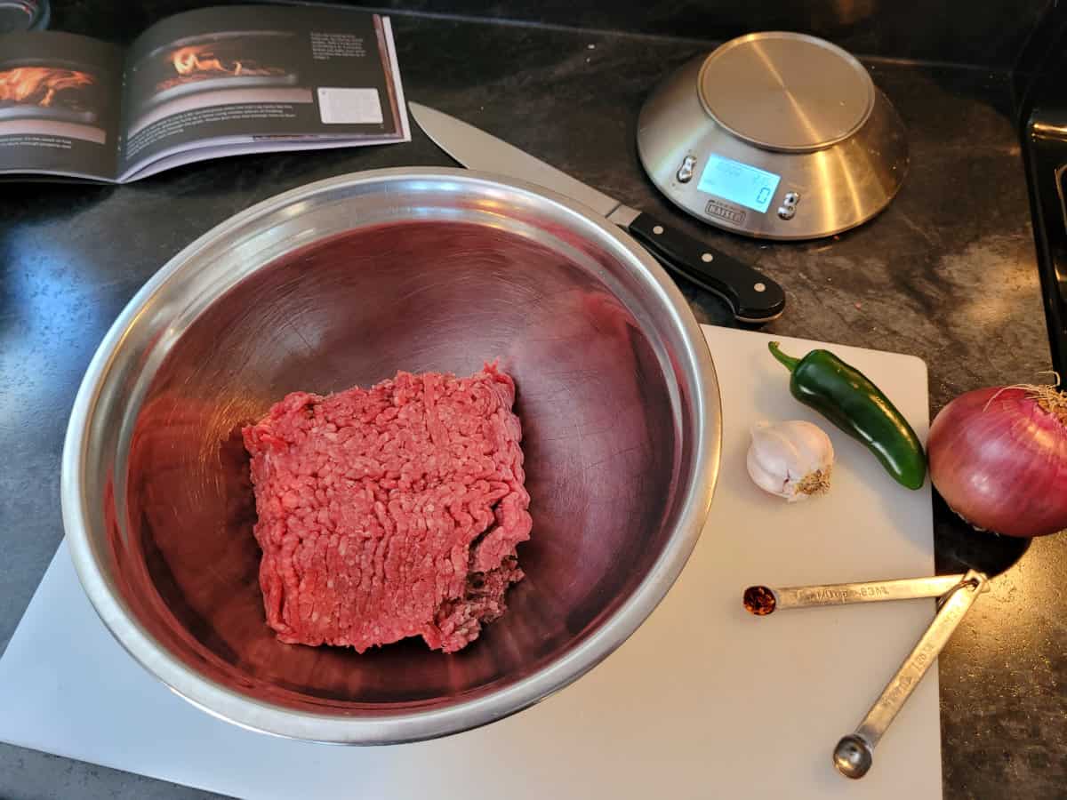 Ground beef and various ingredients needed to make meatballs displayed in a bowl and on a cutting board.