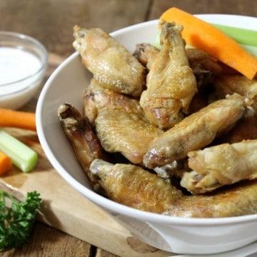 crispy baked chicken wings in a white bowl on a wooden tabletop with various garnishes.