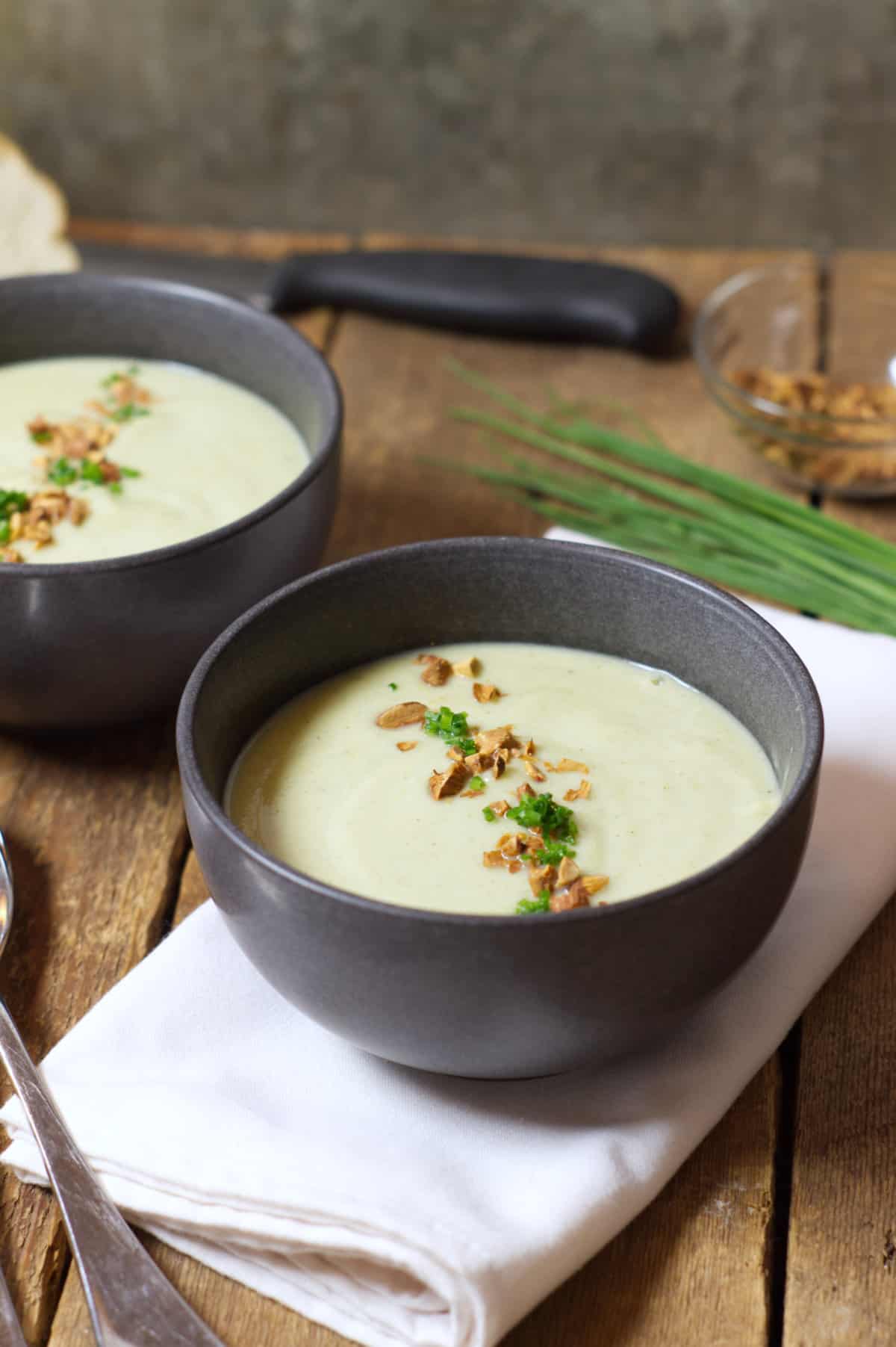 Vertical image of a close up of black stoneware soup bowls filled with pale green broccoli and cauliflower soup and garnished with toasted almonds and chopped chives.