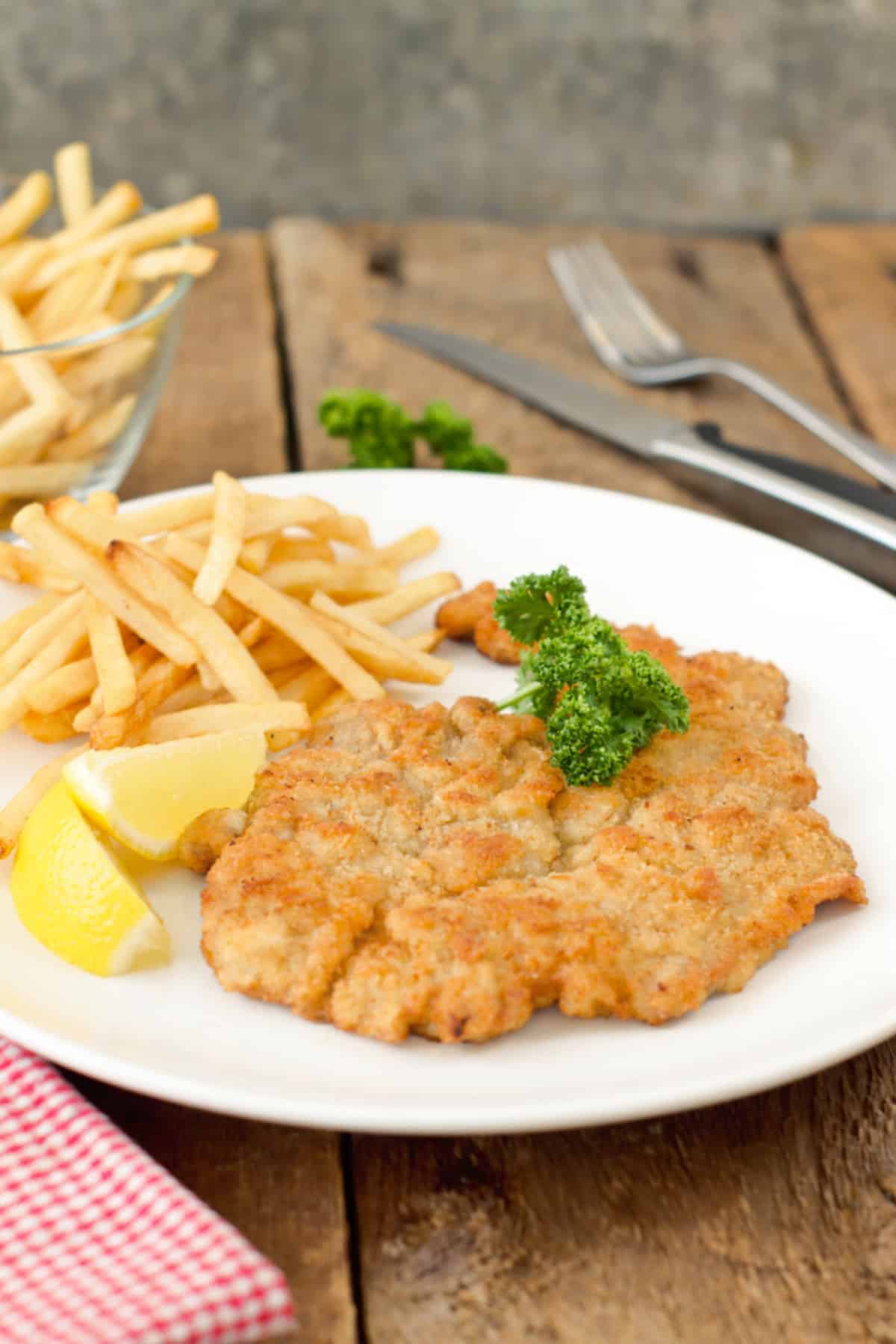vertical image of pork schnitzel being served with french fries and lemon wedges and parsley sprigs on a white plate