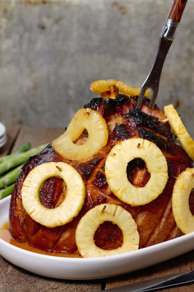 vertical image of a pineapple and honey glazed bone in ham on a white serving platter with a carving fork stuck in it